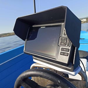 Puede incluir: Un buscador de peces Garmin negro con una visera protectora est&aacute; montado en un barco azul. El dispositivo tiene una pantalla y varios botones. El barco est&aacute; en un lago con un cielo azul claro en el fondo.