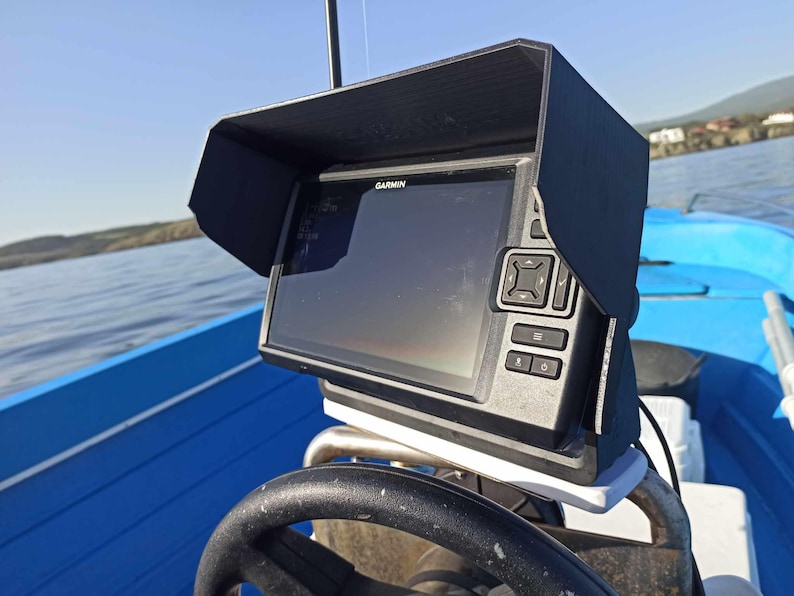 Puede incluir: Un buscador de peces Garmin negro con una visera protectora est&aacute; montado en un barco azul. El dispositivo tiene una pantalla y varios botones. El barco est&aacute; en un lago con un cielo azul claro en el fondo.