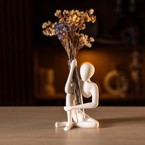 以下が含まれることがあります： 座っている人物の白いセラミック彫刻。透明な花瓶にはドライフラワーが入っています。花は黄色と青です。彫刻は明るい茶色の表面にあります。