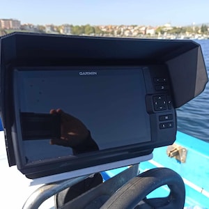 Puede incluir: Un buscador de peces Garmin montado en un barco. El dispositivo tiene una pantalla negra y est&aacute; encerrado en una cubierta protectora negra. La pantalla muestra el logotipo de Garmin. El barco est&aacute; en el agua con una ciudad al fondo.