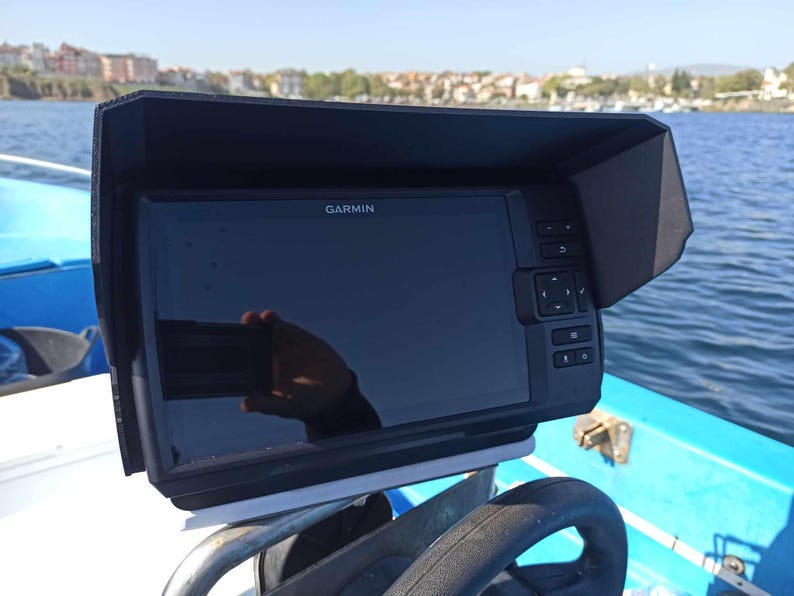Puede incluir: Un buscador de peces Garmin montado en un barco. El dispositivo tiene una pantalla negra y est&aacute; encerrado en una cubierta protectora negra. La pantalla muestra el logotipo de Garmin. El barco est&aacute; en el agua con una ciudad al fondo.