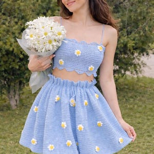 Op de afbeelding: Een lichtblauwe gehaakte tweedelige set met madeliefjesaccenten. De set bestaat uit een crop top met spaghettibandjes, een rok met hoge taille en een bijpassende hoofdband. De outfit is versierd met witte en gele madeliefjesapplicaties.