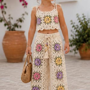 Puede incluir: Un conjunto de dos piezas de ganchillo compuesto por un top corto con tirantes anudados y pantalones anchos. Ambas prendas están hechas de hilo color crema con motivos florales cuadrados en tonos amarillo, rosa y lavanda.