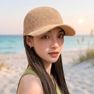 May include: A beige, woven baseball cap with a curved peak. The cap is made of a light, breathable material, ideal for sun protection. The cap is worn by a person with long, dark hair and a green top, with a beach background.