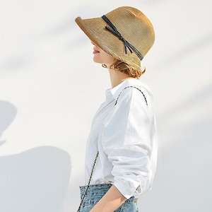 Può includere: Un cappello da sole di paglia marrone chiaro con tesa larga e nastro blu scuro. Il cappello è indossato da una persona che indossa una camicia bianca e jeans blu. Il cappello ha una forma leggermente curva.