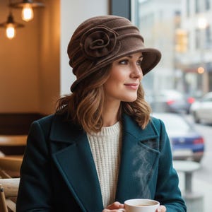 May include: A brown cloche hat with a large flower detail. The hat has a small brim and is made of a soft material. The woman is wearing a teal coat and a cream-colored sweater.