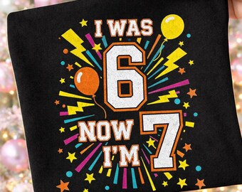以下が含まれることがあります： I WAS 6 NOW I'M 7というテキストが書かれたカラフルな誕生日グラフィックが特徴の黒いTシャツ。デザインには、オレンジ色の輪郭を持つ大きな白い数字の6と7が含まれており、黄色の稲妻、星、オレンジと黄色の風船で囲まれています。