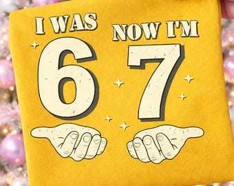以下が含まれることがあります： マスタードイエローのTシャツで、太字のヴィンテージ風フォントで「I WAS 6 NOW I'M 7」というテキストがプリントされています。数字の下には開いた手のイラストが2つ描かれ、生地の表面には小さな星のアクセントが散りばめられています。