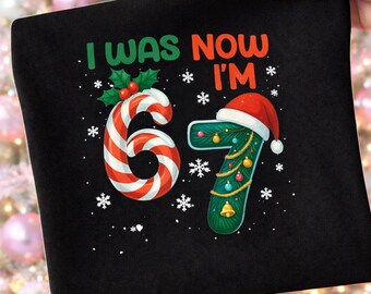 以下が含まれることがあります： 緑とオレンジで「I WAS NOW I'M 67」と書かれた黒い布製品。数字の6はヒイラギの葉が付いた赤と白のキャンディケインで、数字の7はオーナメントとサンタの帽子で飾られた緑色のクリスマスツリーのデザインです。