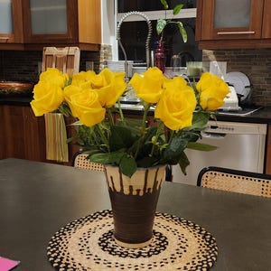 Puede incluir: Un ramo de rosas amarillas vibrantes en un jarrón decorativo con diseño marrón y crema. El jarrón está sobre un salvamanteles redondo y tejido con borde negro y crema. Sobre una mesa gris oscura.
