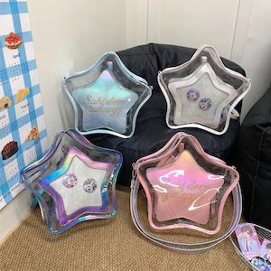Puede incluir: Cuatro bolsos cruzados en forma de estrella con exteriores transparentes e iridiscentes. Cada bolso tiene un interior de color pastel diferente: azul, blanco y rosa. El texto "Saki deers galaxy" está impreso en la parte delantera de los bolsos. Algunos bolsos tienen alfileres decorativos.