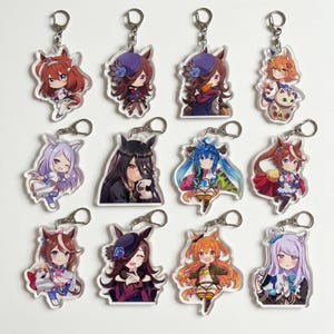 Könnte beinhalten: Eine Sammlung von zwölf Schlüsselanhängern im Anime-Stil, die verschiedene Charaktere mit pferdeähnlichen Ohren und Schwänzen zeigen. Jeder Schlüsselanhänger zeigt einen anderen Charakter mit einzigartigen Outfits und Frisuren. Die Schlüsselanhänger sind aus klarem Acryl gefertigt und haben silberne Schlüsselringe.