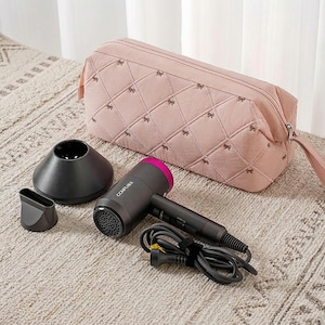 May include: A black and pink hair dryer with attachments and a matching pink quilted cosmetic bag. The bag is decorated with bow patterns. The hair dryer has a black handle and a pink accent on the nozzle. The set is on a neutral-colored rug.
