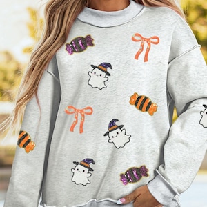 Puede incluir: Una sudadera gris claro de manga larga con cuello redondo y dobladillo de borde sin rematar. La prenda está decorada con parches de lentejuelas dispersos de fantasmas blancos con sombreros morados y naranjas, lazos naranjas y caramelos.