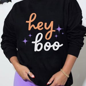 Puede incluir: Una sudadera negra de manga larga con el texto "hey" en naranja y "boo" en blanco, acentuado con pequeños gráficos de estrellas moradas de cuatro puntas. La prenda tiene cuello redondo y puños acanalados, ofreciendo un ajuste estándar.
