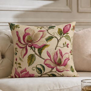May include: A cream-colored sofa with a floral patterned pillow featuring pink magnolia blossoms. A crystal decanter and a glass of amber liquid sit on a gold tray on a marble table. A framed botanical print hangs on the wall.