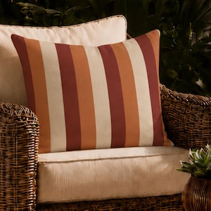 May include: A square throw pillow featuring vertical stripes in alternating shades of cream, burnt orange, and deep red. The pillow is positioned on a woven wicker armchair with a light-colored seat cushion, set in an outdoor environment.