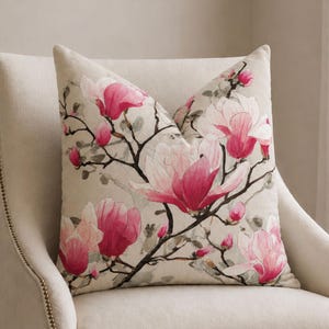 Può includere: Un cuscino decorativo con un design floreale di magnolia. Il cuscino presenta grandi fiori di magnolia rosa e bianchi su uno sfondo beige. Il cuscino è posto su una sedia imbottita color crema.