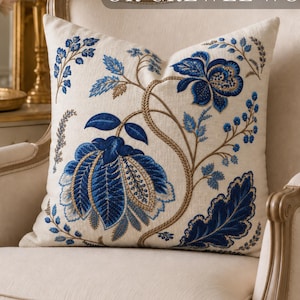 Pode incluir: Uma almofada quadrada com uma base de cor creme apresentando uma estampa botânica floral em azul e dourado. O texto PRINTED NOT EMBROIDERED OR CREWEL WORK é visível no topo. O design exibe grandes flores e folhas azuis.