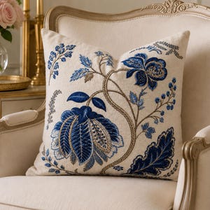 May include: A square throw cushion featuring an off-white fabric base with intricate embroidery in shades of deep blue, light blue, and metallic gold. The design displays a large floral motif with winding stems, leaves, and small berry clusters.