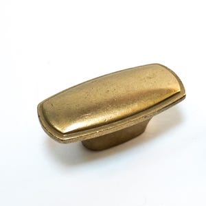 May include: Two cabinet knobs. One is a rectangular shape with a brass finish, and the other is a similar shape with a black finish. Both knobs are on a white background.