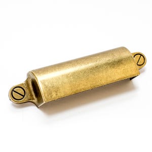 May include: A brass-colored, rectangular cabinet pull with rounded ends and two visible screws. The hardware has a slightly aged appearance, suggesting a vintage or antique style. The pull is designed for easy gripping and opening of drawers or cabinets.