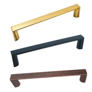 May include: Three cabinet pulls in gold, black, and bronze. Each pull features a rectangular bar with curved ends. The gold pull has a slightly distressed look, while the black and bronze pulls have a smooth finish. Ideal for kitchen or furniture upgrades.