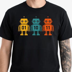 May include: Black t-shirt featuring three colorful robot illustrations. Each robot is a different color: yellow, teal, and orange. The robots have the numbers 01, 10, and 11 on their chests. The robots have a retro, cartoon style.