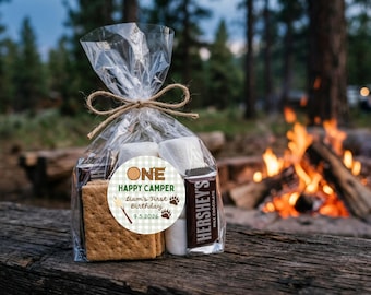 Könnte beinhalten: Ein durchsichtiger Plastikbeutel mit Bindfaden enthält Graham-Cracker, Marshmallows und eine Hershey's Milchschokoladentafel. Ein runder Aufkleber auf der Vorderseite zeigt den Text ONE HAPPY CAMPER Liam's First Birthday 9.5.2026 mit Bärentatzen.