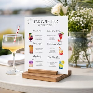 Può includere: Un cartello rettangolare bianco intitolato Lemonade Bar Recipe Ideas, tenuto da un supporto in legno. Il cartello presenta sei illustrazioni ad acquerello di cocktail con nomi come Berry Proud e Blue Skies Ahead, elencando gli ingredienti.