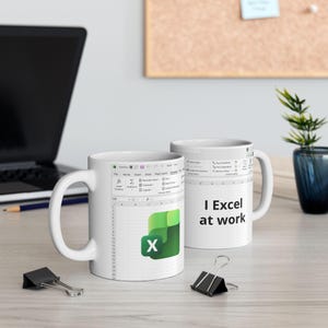 May include: Two white ceramic mugs with black handles. One mug features the Microsoft Excel logo and spreadsheet interface. The other mug has the text "I Excel at work" in black. The mugs are on a light-colored surface.