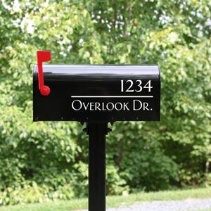 May include: A black metal mailbox mounted on a post featuring a bright red signal flag on the left side. The front of the mailbox displays the white text 1234 above a horizontal line and the street name Overlook Dr. in a clean white font.