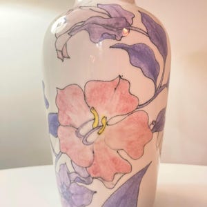 May include: A white ceramic vase with a floral design. The vase features large pink flowers with yellow centers and purple leaves and stems. The flowers are hand-painted on the vase, creating a unique and artistic look. The vase has a smooth, glossy finish.