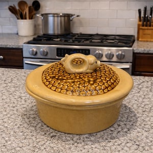 May include: A tan ceramic casserole dish with a lid featuring a textured pattern and a pig-shaped handle. The dish is on a speckled countertop in a kitchen setting. The lid has a brown and tan pattern.