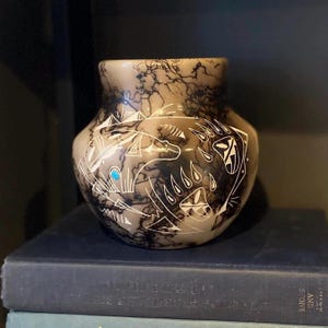 Puede incluir: Un jarrón de cerámica de color beige con un patrón de relámpagos negro y diseños geométricos blancos. El jarrón presenta un acento turquesa y un diseño de pata de oso. El jarrón está sobre una pila de libros.