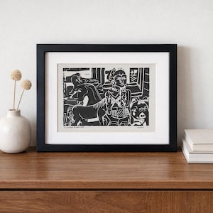 May include: A framed black and white print featuring a stylized illustration of a person, a horse, and a dog. The artwork is in a black frame and is displayed on a wooden surface. A vase with dried flowers is to the left.