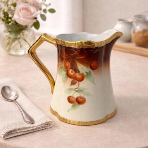 May include: A ceramic pitcher with a gold handle and trim. The pitcher has a hand-painted design of cherries and green leaves. The top of the pitcher is white, transitioning to brown, and then to white.