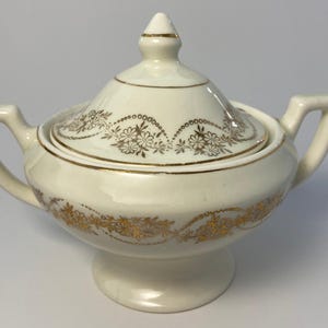 Puede incluir: Azucarero crema con tapa y detalles florales dorados. El cuenco tiene dos asas y se apoya sobre una pequeña base. La tapa está rematada con un pequeño adorno. El diseño floral dorado se repite alrededor del cuenco y la tapa.