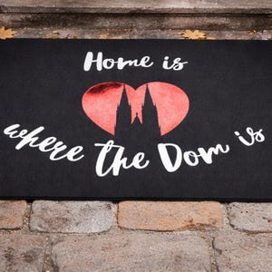 Könnte beinhalten: Schwarze Fußmatte mit dem weißen Schriftzug "Home is where the Dom is". Ein rotes Herz mit einer Silhouette einer Kathedrale befindet sich in der Mitte. Braune Stiefel und eine Laterne mit brennender Kerze links.