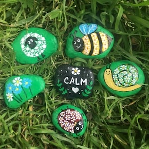 Puede incluir: Rocas pintadas a mano con varios diseños, incluyendo una abeja, una mariquita, un caracol y flores, sobre un fondo de hierba verde. Una roca tiene la palabra "CALM" pintada. Las rocas están pintadas con colores brillantes.