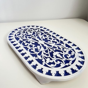 May include: An oval decorative tray in white and blue. The tray has a white, openwork floral design set against a blue background. The border is blue with white leaf-like cutouts. It has a small, white foot. Measurements are approximately 30 cm long.