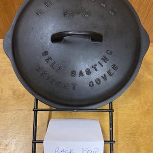 May include: A black cast iron Griswold No. 8 self-basting skillet cover. The lid has a handle and is displayed on a black metal rack. The text "SELF BASTING COVER" and "GRISWOLD No 8" are embossed on the lid.