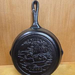 May include: A black cast iron skillet featuring a raised relief design on the bottom depicting a duck swimming in a pond with reeds. The word OTTER is embossed at the bottom of the design. The skillet has a long handle with a hole for hanging.