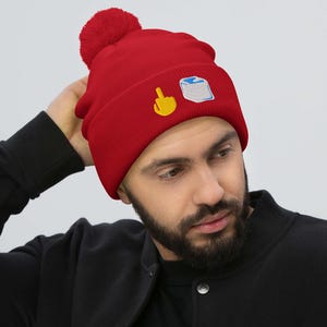 May include: Red beanie with a matching pom-pom. The beanie features embroidered designs of a yellow middle finger and a white and blue square. The person in the image is wearing a black jacket.