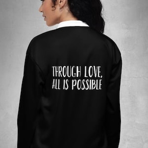 May include: Black bomber jacket with white trim and the text "THROUGH LOVE, ALL IS POSSIBLE" in white. The jacket has a white collar and cuffs. The Midnight Thread Shop logo is on the lower right.