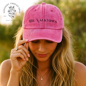 May include: A pink baseball cap with the text "THE VALKYRIES" in black. The cap is worn by a person with long blonde hair. The Midnight Thread Shop logo is visible on the left side of the image.