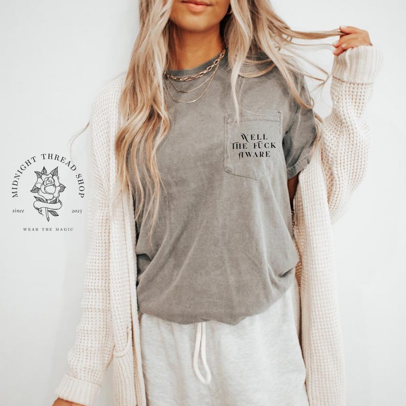 May include: A gray pocket t-shirt with the text "Well the fuck aware" printed on the pocket. The shirt is part of a casual outfit, paired with gray sweatpants and a cream-colored cardigan. The Midnight Thread Shop logo is visible.
