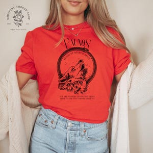 May include: A red t-shirt with a black graphic of a howling wolf inside a circular frame. The text "FAUNOS" is above the wolf, with additional text below. The shirt is part of the "Midnight Thread Shop" collection.