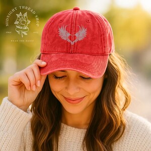 May include: Red baseball cap with a heart and wings design. The cap has a curved brim and a distressed look. The text "Midnight Thread Shop" and "Wear the Magic" are also visible.