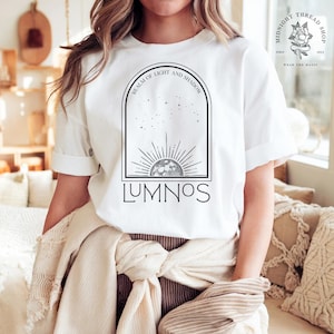 May include: White t-shirt with a black graphic design featuring a sun and moon illustration, with the text "REALM OF LIGHT AND SHADOW" above and "LUMNOS" below. The shirt has rolled-up sleeves and a relaxed fit.